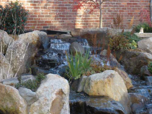natural stone backyard waterfall flowing through landscaped pond area beside brick wall